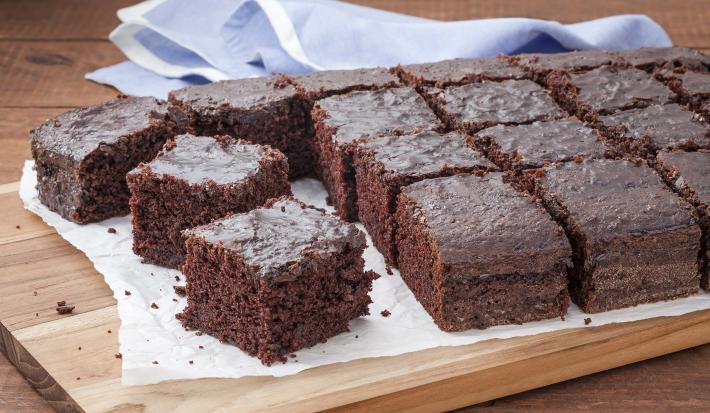 16 pedaços de bolo de chocolate com calda em cima de uma toalha branca que está em cima de uma tábua de madeira. Todos do mesmo tamanho