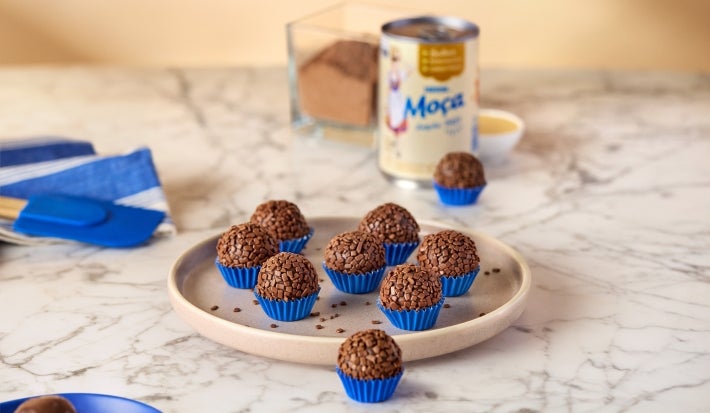 Fotografia mostra brigadeiros enrolados com granulado em forminhas azuis. No fundo, a lata de leite moça.
