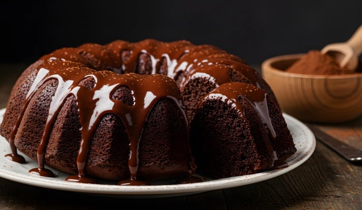 A imagem apresenta um bolo de chocolate Bundt, disposto em um prato cerâmico branco. A cobertura de chocolate brilhante escorre suavemente pelas laterais do bolo. A textura do bolo parece densa e úmida.
