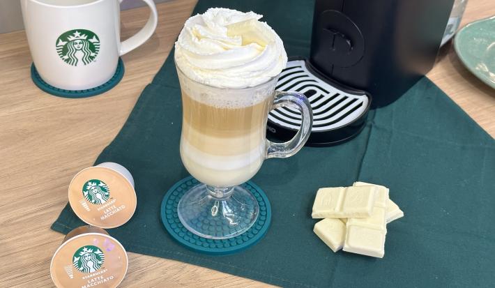 Foto da receita de Mocha Branco. Observa-se uma xícara transparente com a bebida em fases decorada com chantilly e, ao lado, cápsulas de Starbucks e quadradinhos de Galak.