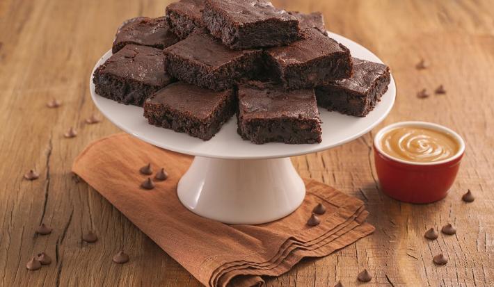 Fotografia em tons de marrom e vermelho de uma bancada de madeira com um paninho, sobre ele um prato branco com pedaços de brownie. Ao lado um potinho com Moça de colher doce de leite.