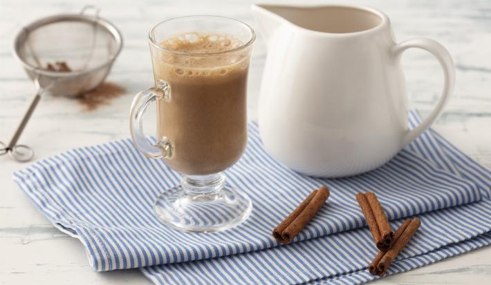 Fotografia em tons de azul em uma bancada de madeira clara com um pano azul com listras finas e ao centro uma taça com o cappuccino e ao lado uma jarrinha branca. Ao lado, três paus de canela e um difusor com o chocolate em pó.