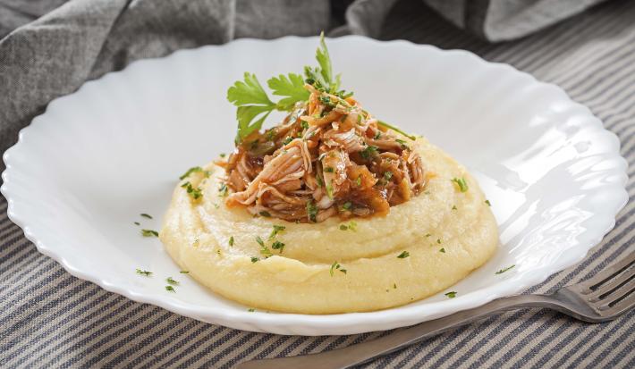 Fotografia em tons de cinza, branco e amarelo, prato branco ao centro com polenta, frango desfiado e salsa, ao lado um garfo base, sobre toalha com listras brancas e cinza e ao fundo guardanapo cinza.