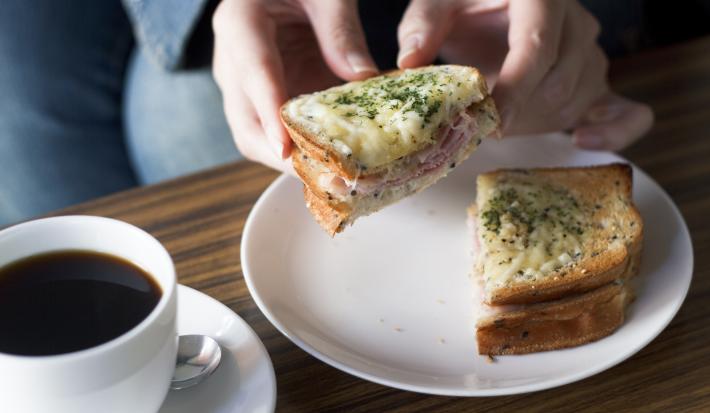 Fotografia de um sanduíche aberto com uma moça segurando a metade do lanche, que tem presunto, lombinho defumado, molho e queijo gruyère gratinado por cima. O sanduíche está em um prato raso branco de vidro ao lado de uma xícara branca de café cheia.