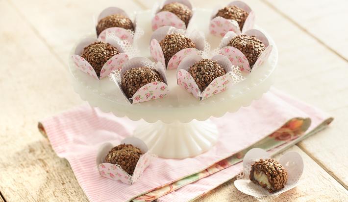 fotografia em tons de cinza, rosa e marrom de uma bancada cinza vista de cima, contém um suporte branco para servir sobremesas com forminhas rosas com brigadeiros dentro, e abaixo um pano rosa com 2 forminhas e ambas contém brigadeiro