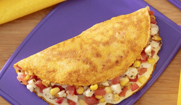 Fotografia em tons de roxo com um prato roxo ao centro. Em cima do prato existe uma receita de crepioca recheada com tomate, milho e palmito.