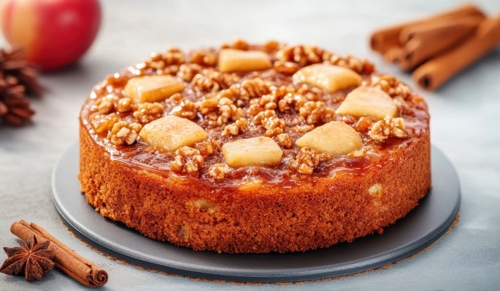 Fotografia de um bolo de maçã e nozes, com crosta dourada e cobertura de caramelo, decorado com pedaços de maçã e nozes inteiras, em um prato escuro, cercado por canela e anis estrelado.