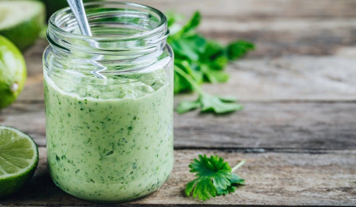 Fotografia mostra um pote transparente com uma maionese verde dentro. O pote está sobre uma mesa de madeira, ao lado rodelas de limão.