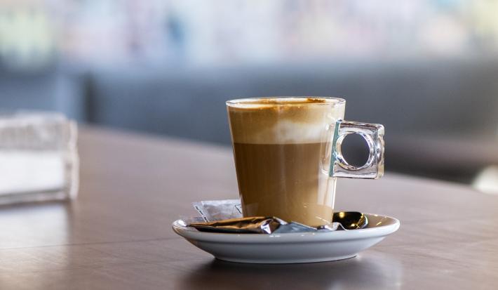 fotografia em tons de marrom e branco de uma bancada marrom vista de frente. Contém um pratinho redondo branco com uma xicara de café transparente, dentro contém café e chantilly por cima.