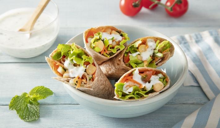 Foto em tons coloridos da receita de wrap vegetariano com molho de hortelã servida em 4 porções dentro de um bowl de porcelana branca sobre uma mesa de madeira azul clara com um paninho listrado azul e um pote de iogurte natural