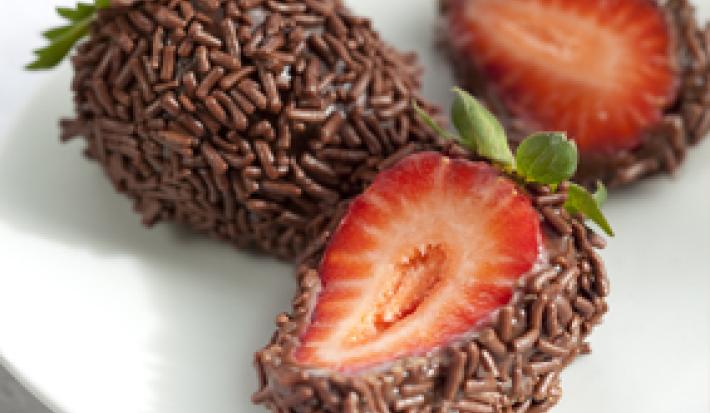 fotografia em tons de branco, marrom e vermelho tirada de um prato branco, contém 3 morangos cobertos por chocolate e chocolate granulado. Dois deles estão pela metade mostrando o morango