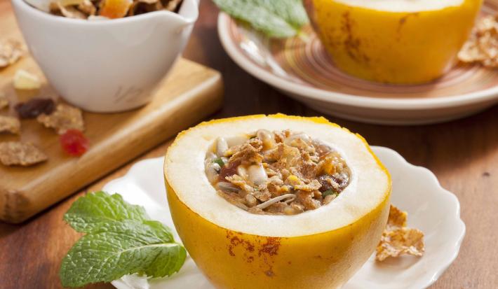 Fotografia em tons de amarelo em uma bancada de madeira, pratos brancos, bege e tábua de madeira cada um com um maracujá doce aberto recheado com Nesfti Cereal Matina com frutas.