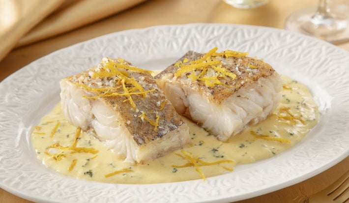 Filés de bacalhau servidos com molho cremoso de ervas e raspas de limão, apresentados em prato branco elegante.