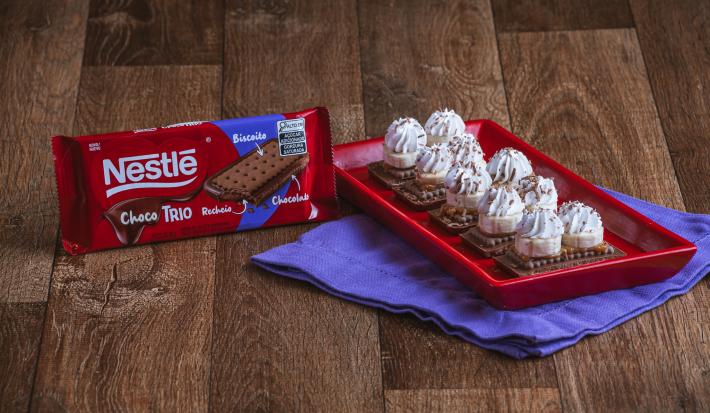Fotografia de pedaços de choco trio chocolate sobre um prato retangular vermelho, cobertos por rodelas de banana, doce de leite e chantilly.