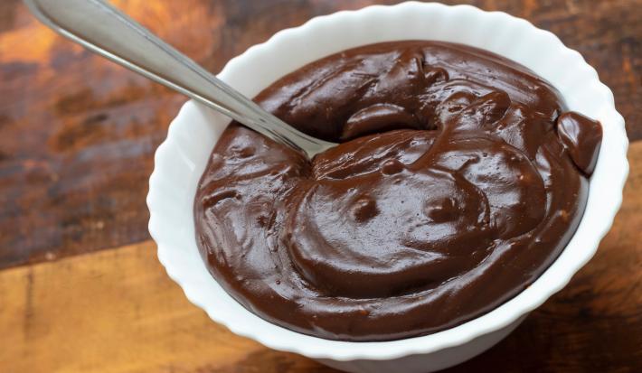 Fotografia de um recipiente pequeno branco fundo com brigadeiro de whey. O recipiente está sobre uma mesa de madeira.