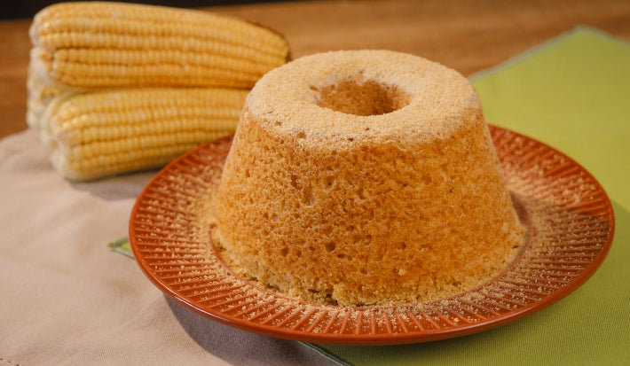 Imagem mostra bolo de milho redondo com furo no meio em um prato vermelho e ao lado de de espigas de milho
