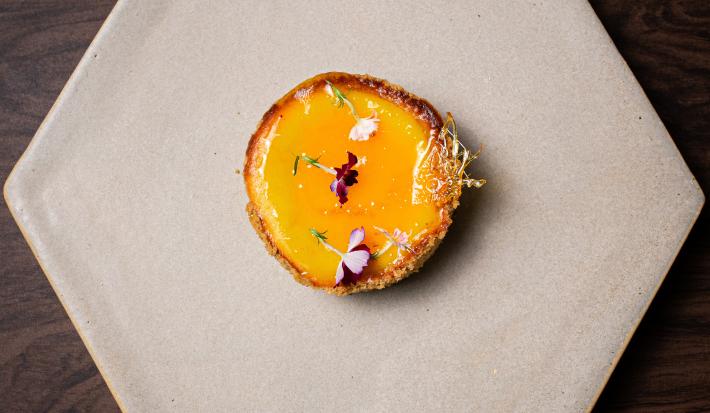 Fotografia em tons de cinza com uma cheesecake amarela ao centro. Por cima da cheesecake existe uma calda de bergamota, caramelo e flores comestíveis.