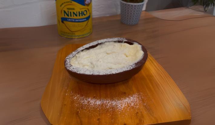 Fotografia em tons de marrom claro. No centro da imagem existe uma casca de ovo recheada com uma mousse de leite NINHO e polvilhada com leite NINHO. Ao fundo existe uma lata de leite NINHO.