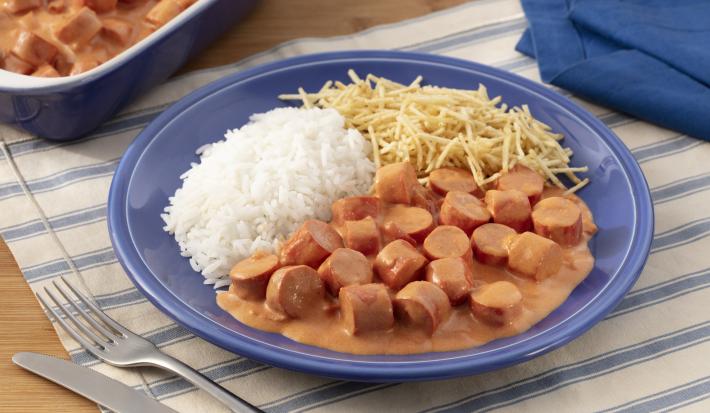 Fotografia em tons de azul em uma bancada de madeira com um jogo americano branco com listras azuis, um prato fundo azul com o strogonoff de salsicha, arroz branco e batata palha. Ao fundo, um recipiente com mais strogonoff de salsicha.