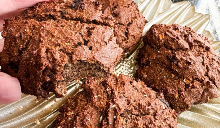 Fotografia em tons claros de uma mão feminina segurnaod um cookie fit, grande, de nescau. No fundo tem um prato raso dourado com mais unidades do cookie.