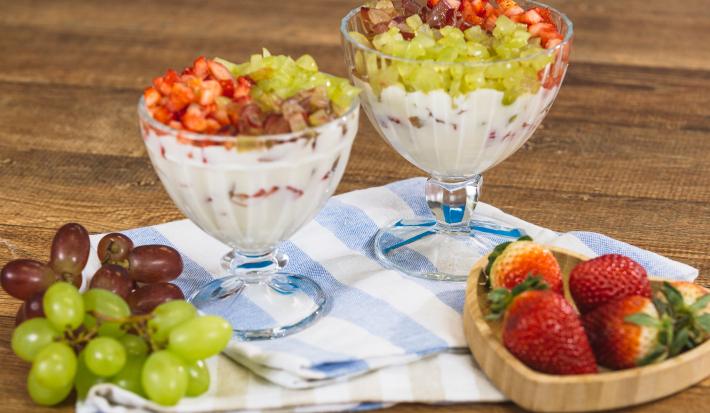 Fotografia em tons de azul e bege em uma bancada e madeira escura, um pano listrado em azul e bege, duas taças de vidro com a sobremesa de creme com morangos e uvas picados. Ao lado, um potinho com morangos em cima e cachos de uvas.