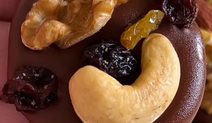 Fotografia em tons de marrom com várias moedinhas de chocolate ao centro enfeitadas com frutas secas, nozes e castanha de caju