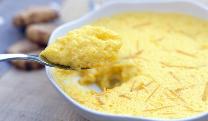 Fotografia em tons claros de uma mousse de acerola em um recipiente branco fundo. No centro, uma colher carregando um pouco da mousse, que está sobre uma toalha de mesa, e ao fundo, alguns biscoitos.