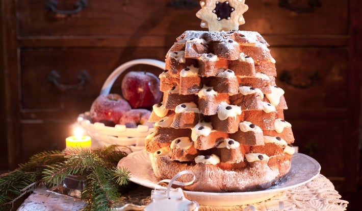Em cima de uma mesa, panetone decorado com creme branco em formato de árvore. No topo do panetone, biscoito em formato de estrela.