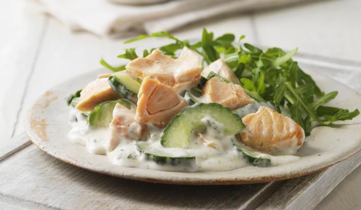 Fotografia de um pepino refrescante e cremoso com salmão defumado e uma salada verde em um prato raso de cerâmica de cor clara. O prato está sobre uma tábua de madeira branca, apoiada em uma mesa de madeira do mesmo tom.