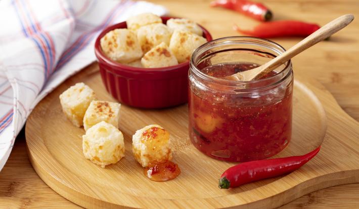 Fotografia em tons de roxo em uma bancada de madeira clara, um pano branco com lilás xadrez, uma tábua de madeira clara com um potinho de vidro e a geleia de pimenta dentro. Ao lado, um potinho vermelho com os dadinhos de tapioca.
