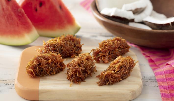 Foto da receita de cocada com casca de melancia. Observa-se uma tábua de madeira com 5 cocadas dispostas. Atrás, uma decoração com pedaços de melancia e pedaços de coco