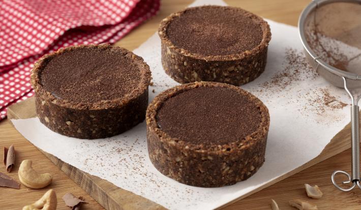 Fotografia em tons de marrom e vermelho de uma bancada de madeira com uma tábua de madeira e uma folha de papel manteiga, sobre ele três tortinhas de ganache. Ao fundo um paninho xadrez vermelho e branco e uma peneira. A frente castanhas-de-caju.