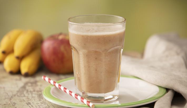 Fotografia em tons de cinza de uma bancada cinza com um prato branco com borda verde sobre ele um canudo listrado branco e vermelho e um copo de vidro com a vitamina de banana e maçã. Ao fundo um cacho de banana, uma maçã e um paninho cinza.