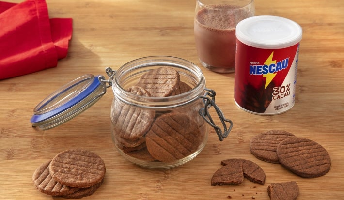 Fotografia de um potinho de vidro com tampa sobre uma mesa de madeira clara, ao lado da lata de NESCAU Extra Cacau 30% e um copo de vidro com leite e nescau. Dentro e em volta do pote tem os biscoitinhos amanteigados de nescau.