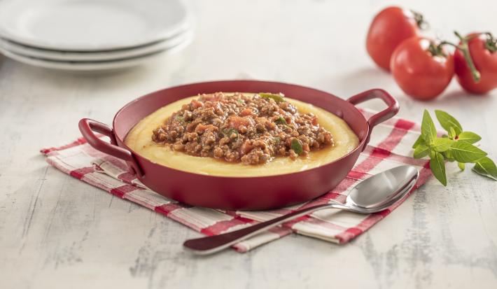 Imagem de uma caçarola vermelha com polenta e carne moída sobre um paninho xadrez vermelho e branco. Compondo a imagem, uma colher, manjericão, tomates e pratos brancos.