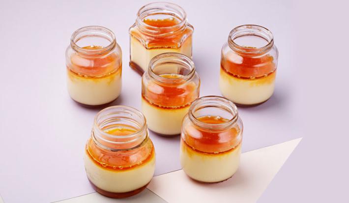 fotografia em tons de branco e laranja vista de cima, contém 6 potinhos transparentes com pudim de dentro e cobertura de caramelo por cima