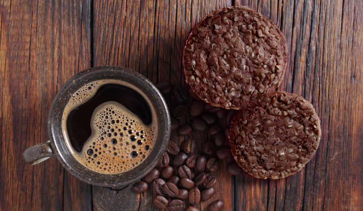 Foto vista de cima da receita de cookies de aveia e café, servidos em uma bancada de madeira, ao lado de uma xícara de café preparada com alguns grãos de café espalhados