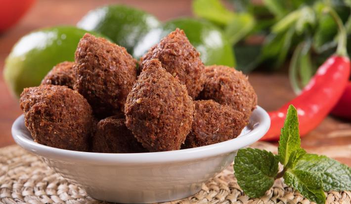 Fotografia em tons de verde com um prato fundo branco ao centro. Dentro do prato existe vários mini kibes feitos na Air fryer. Ao fundo existe limões inteiros e pimentas. Ao lado do prato existe um ramo de hortelã