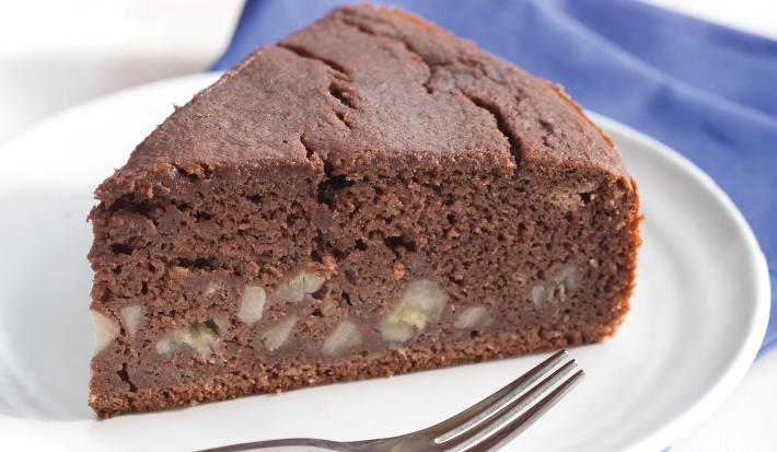 Fotografia em tons de azul em uma mesa de madeira branca com um pano azul, um prato branco raso com uma fatia de bolo de chocolate com banana.