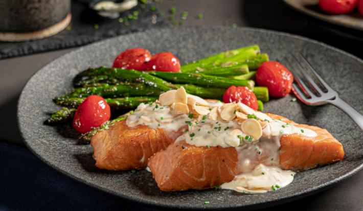 Foto de um prato escuro com a receita de salmão grelhado, acompanhado de aspargos e tomates cereja. Ao fundo há outro prato aparecendo, alguns aspargos um copo preto pequeno com molho dentro