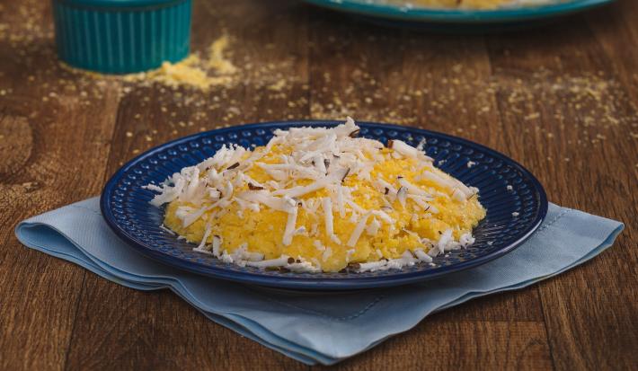 Imagem aproximada da receita de Cuscuz em tom amarelo, em um prato azul escuro, sobre um tecido em tom de azul claro. O cuscuz está decorado com coco ralado e, ao fundo, estão um pote com fubá amarelo e um outro prato com outro cuscuz