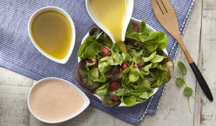 Fotografia em tons de azul, verde e amarelo de uma bancada branca com um paninho azul, sobre ele um prato branco redondo com salada de folhas. Ao redor, três recipiente brancos com molho para salada. Ao lado um garfo para salada.