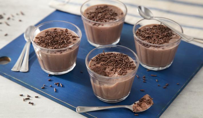 Fotografia em tons de azul em uma bancada de madeira clara, uma tábua azul quadrada com quatro copinhos de vidro com a mousse de chocolate dentro e decoradas com os chocolates granulados.
