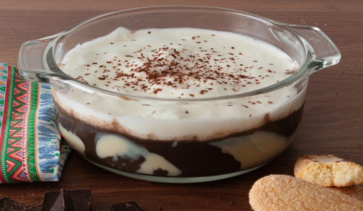 Em cima de uma mesa, recipiente redondo de vidro com creme de chocolate, decorado com chantilly e raspas de chocolate.