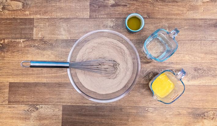Foto vista de cima do recipiente transparente de vidro com os ingredientes secos misturados e, ao lado, os três ingredientes líquidos em potes separados: óleo, água e ovos.