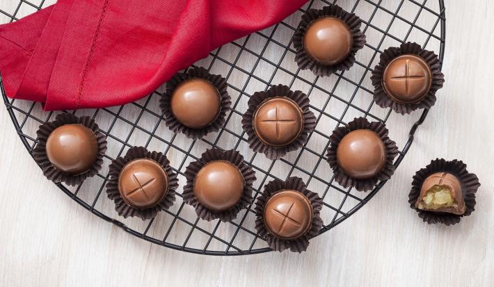 Fotografia em tons de chocolate, branco e vermelho, ao centro vários bombons de chocolate em forminhas de papel marrom sobre grelha redonda, ao lado guardanapo vermelho e bombom aberto, tudo sobre bancada de madeira clara.
