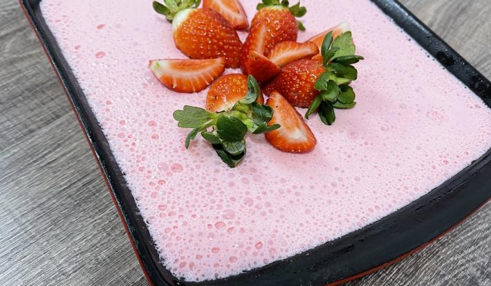 Imagem de uma receita de gelatina em tom rosa, decorada com morangos picados, servida em um refratário preto. Na imagem há um selo com o texto Receita do Consumidor em tom azul claro.