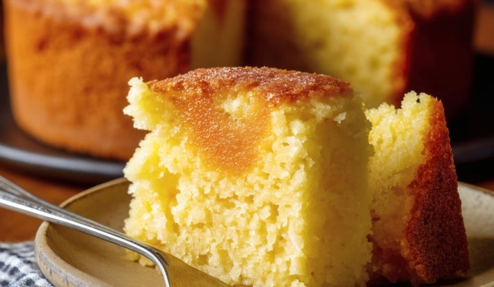 Fotografia de uma fatia de bolo de fubá em um prato cerâmico, mostrando sua textura macia e fofa. O bolo é colocado sobre um prato escuro e está sobre um pano xadrez azul e branco.