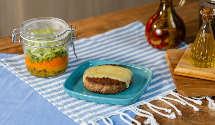 Fotografia em tons de azul com um prato azul ao centro. Em cima do prato existe uma receita de hambúrguer no prato acompanhada por uma fatia de queijo muçarela e uma salada no pote feita com alface e cenoura