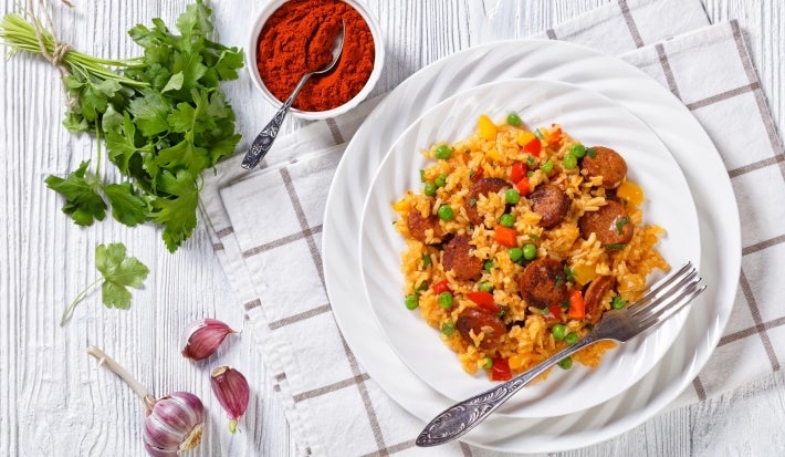 Em cima de uma mesa, visto de cima, prato de porcelana branco com arroz soltinho, calabresa cortada em rodelas, cenouras picadas em cubinhos e ervilhas.